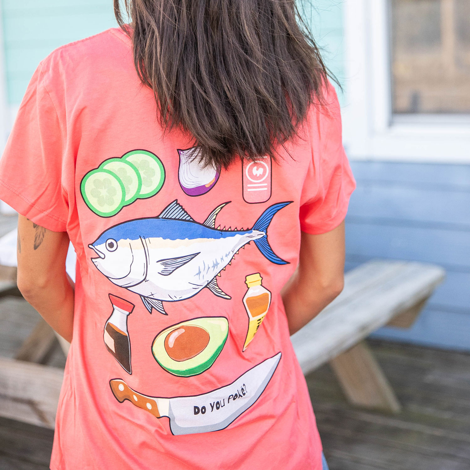 Back of a person wearing a coral t-shirt with illustrations of a fish, avocado, cucumber slices, garlic, sauces, a knife, and the text 'Do you poke?'