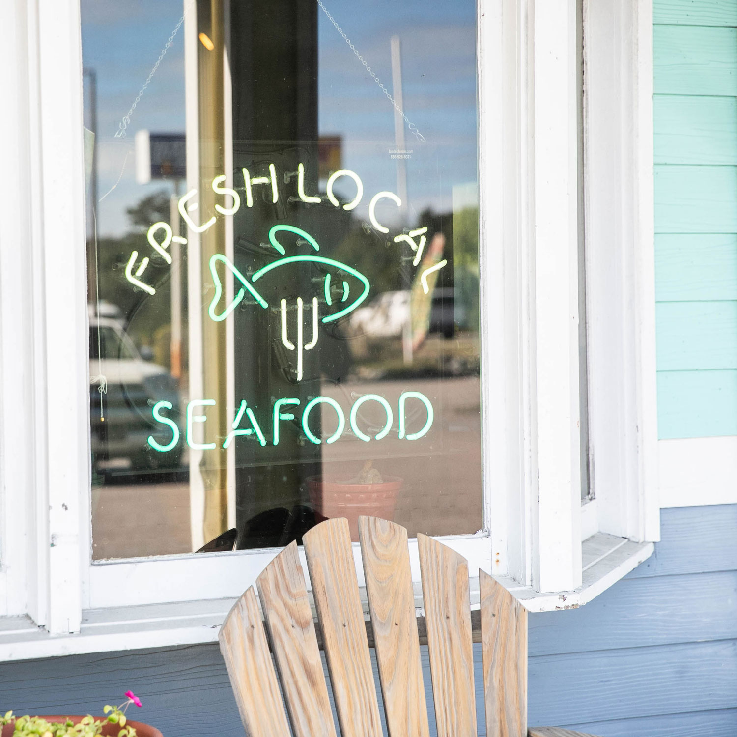 Neon sign reading 'Fresh Local Seafood' with a fish illustration, hanging in a restaurant window above a wooden chair.