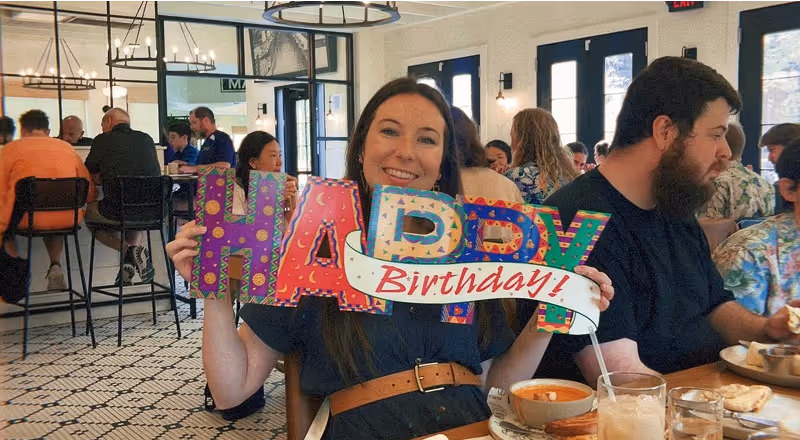 Woman holding a colorful “Happy Birthday” sign while sitting at a restaurant table