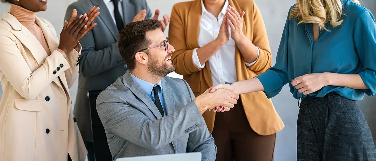 Man in a suit shaking hands with a colleague while team members applaud, representing employee rewards and recognition.