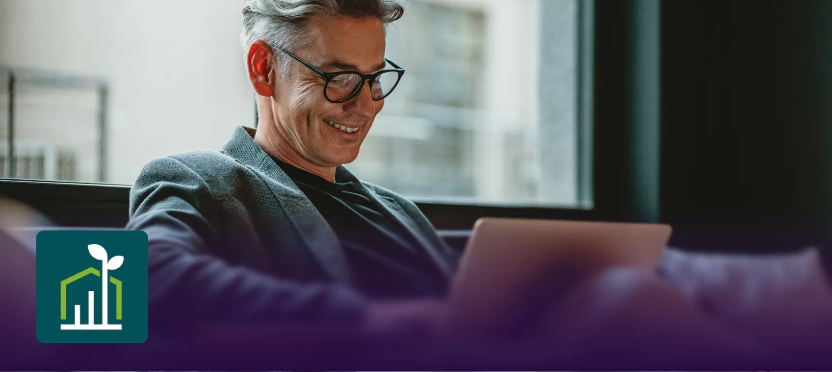 Smiling man wearing glasses and a blazer working on a laptop by a window.