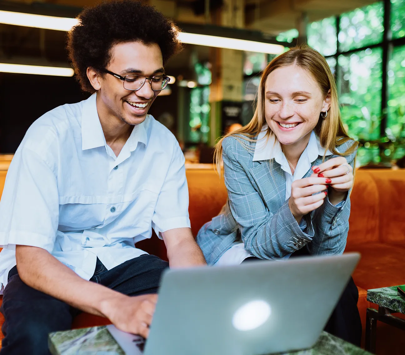 Two young professionals smiling and looking at a laptop while sitting on an orange couch in a bright modern office.