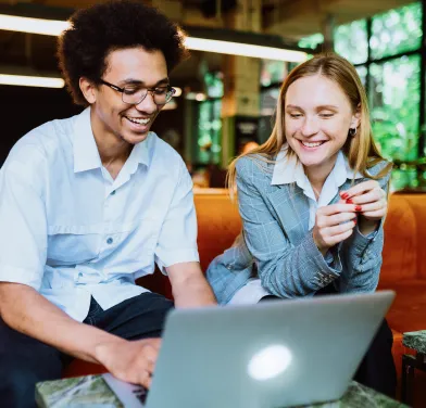 Two smiling young adults sitting on a couch looking at a laptop together in a cozy indoor setting.
