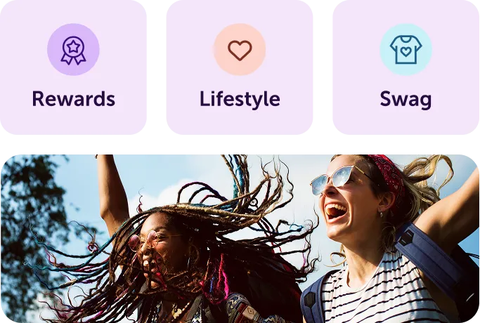 Two joyful women outdoors with wind blowing their hair, smiling and raising their arms under a blue sky.