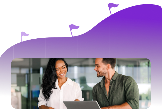 Two coworkers smiling and discussing work in front of a laptop with a purple graph featuring flags in the background.