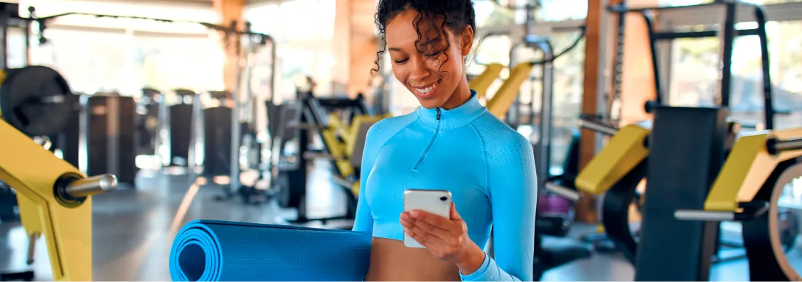 Woman in blue workout clothes holding a yoga mat and looking at her phone in a gym.