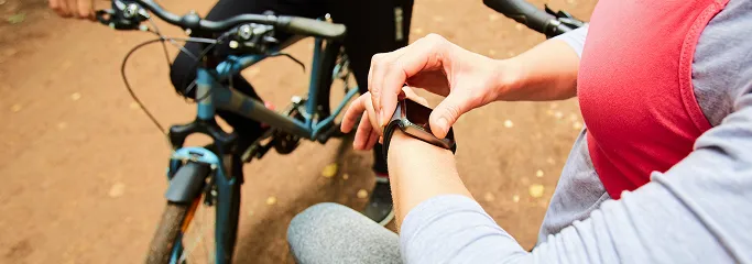 Person wearing a red top adjusts a smartwatch while holding a bicycle on a trail.