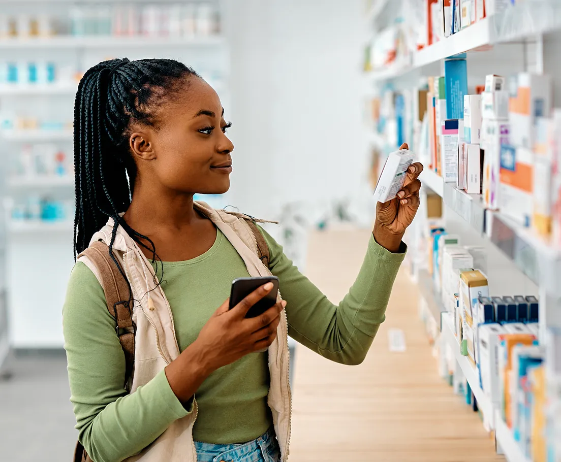 Woman with braided hair holding a phone and reading product packaging in a pharmacy aisle.