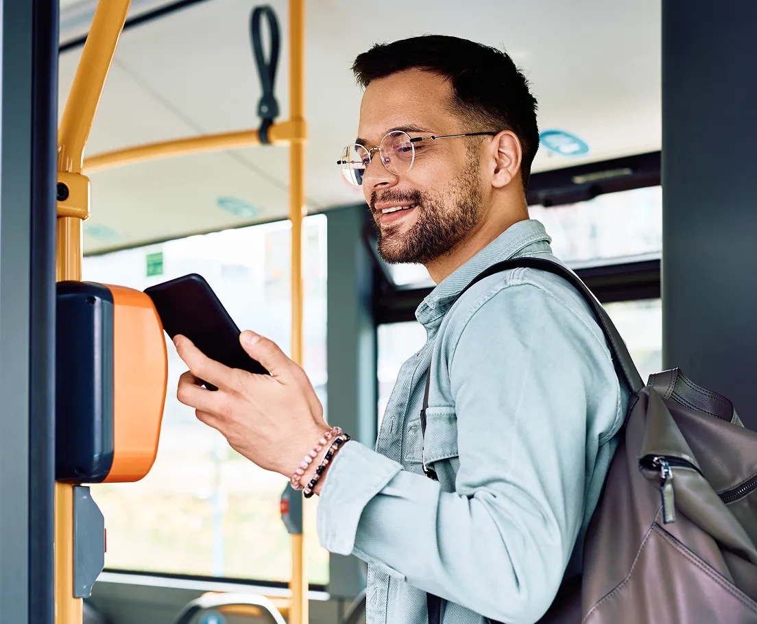 Man with backpack smiling while using smartphone to scan card on bus validation machine.