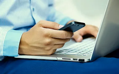 Person in blue shirt holding a smartphone over a laptop keyboard.