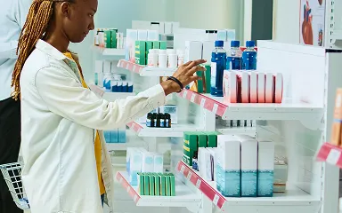Woman in a light-colored coat selecting a product from pharmacy shelves with various medicines and bottles.