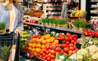 Woman shopping with a cart near fresh tomatoes, greens, and other vegetables in a market.