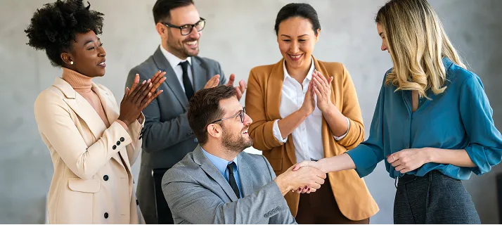 Five colleagues smiling and shaking hands in a workplace setting, with others clapping and happy.