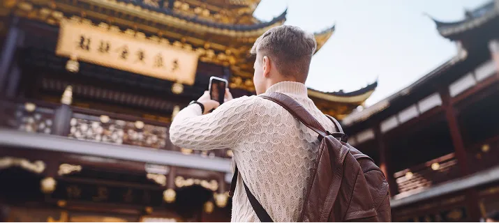 Person in white sweater and brown backpack taking a photo of ornate Asian temple architecture.