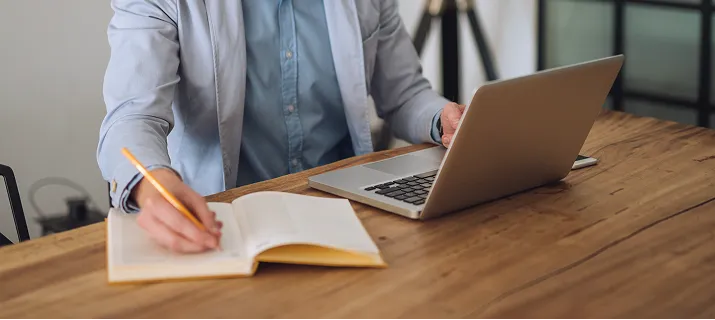 Person in a blazer writing in an open notebook with a pencil while using a laptop on a wooden table.