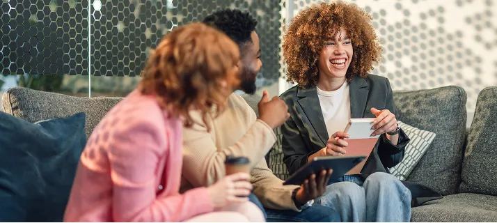 Three friends sitting on a sofa laughing and holding a tablet and color swatches.