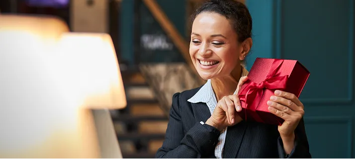 Smiling woman holding a red gift box with a ribbon in a cozy indoor setting.