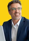 Smiling middle-aged man with glasses wearing a navy suit and white shirt against a yellow background.