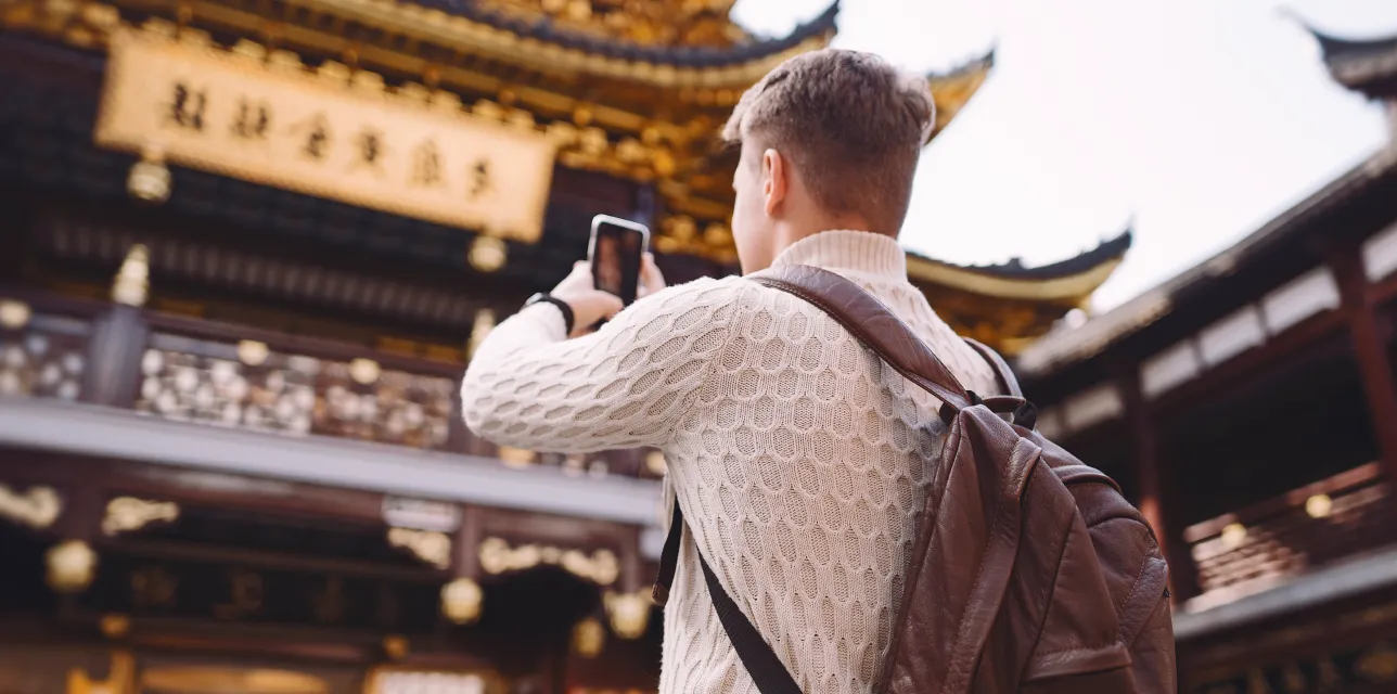 Person in white sweater and brown backpack taking a photo of ornate Asian temple architecture.