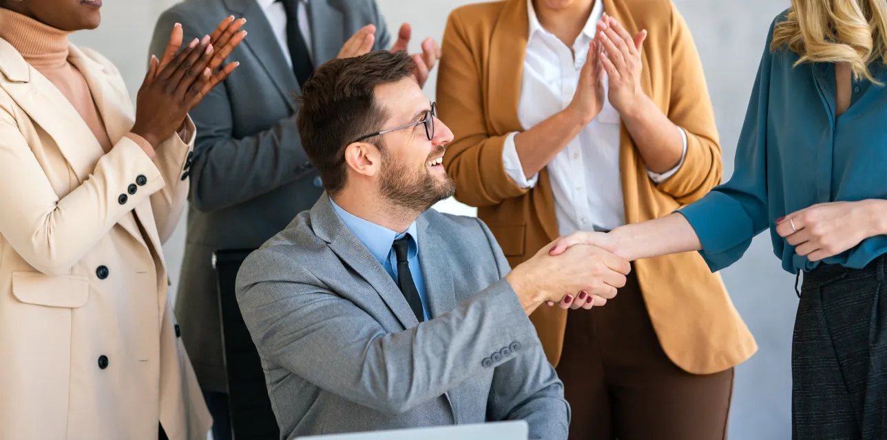 Five colleagues smiling and shaking hands in a workplace setting, with others clapping and happy.