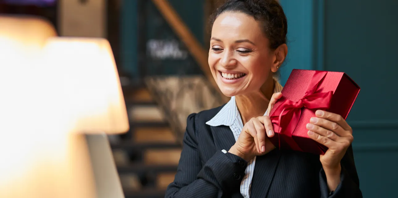 Smiling woman holding a red gift box with a ribbon in a cozy indoor setting.