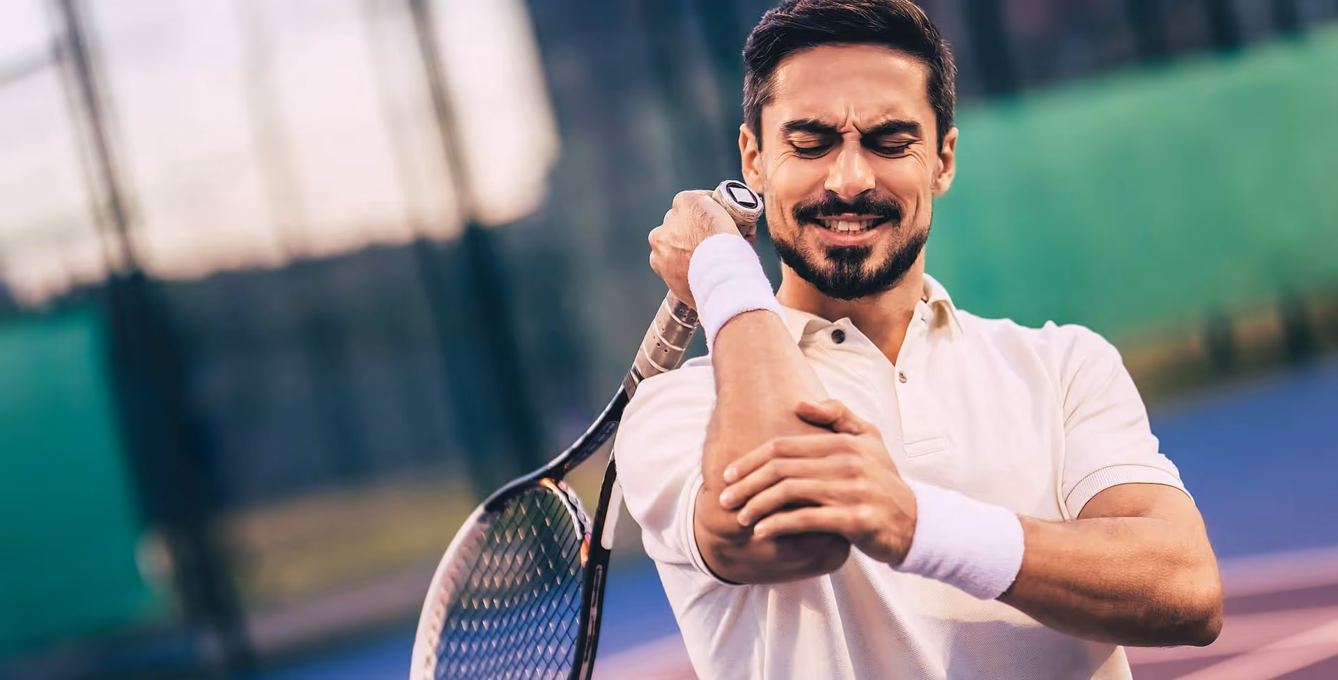 Mann auf einem Tennisplatz, der sich vor Schmerzen den Ellbogen hält, während er einen Schläger umklammert, und Symptome eines Tennisarms zeigt.