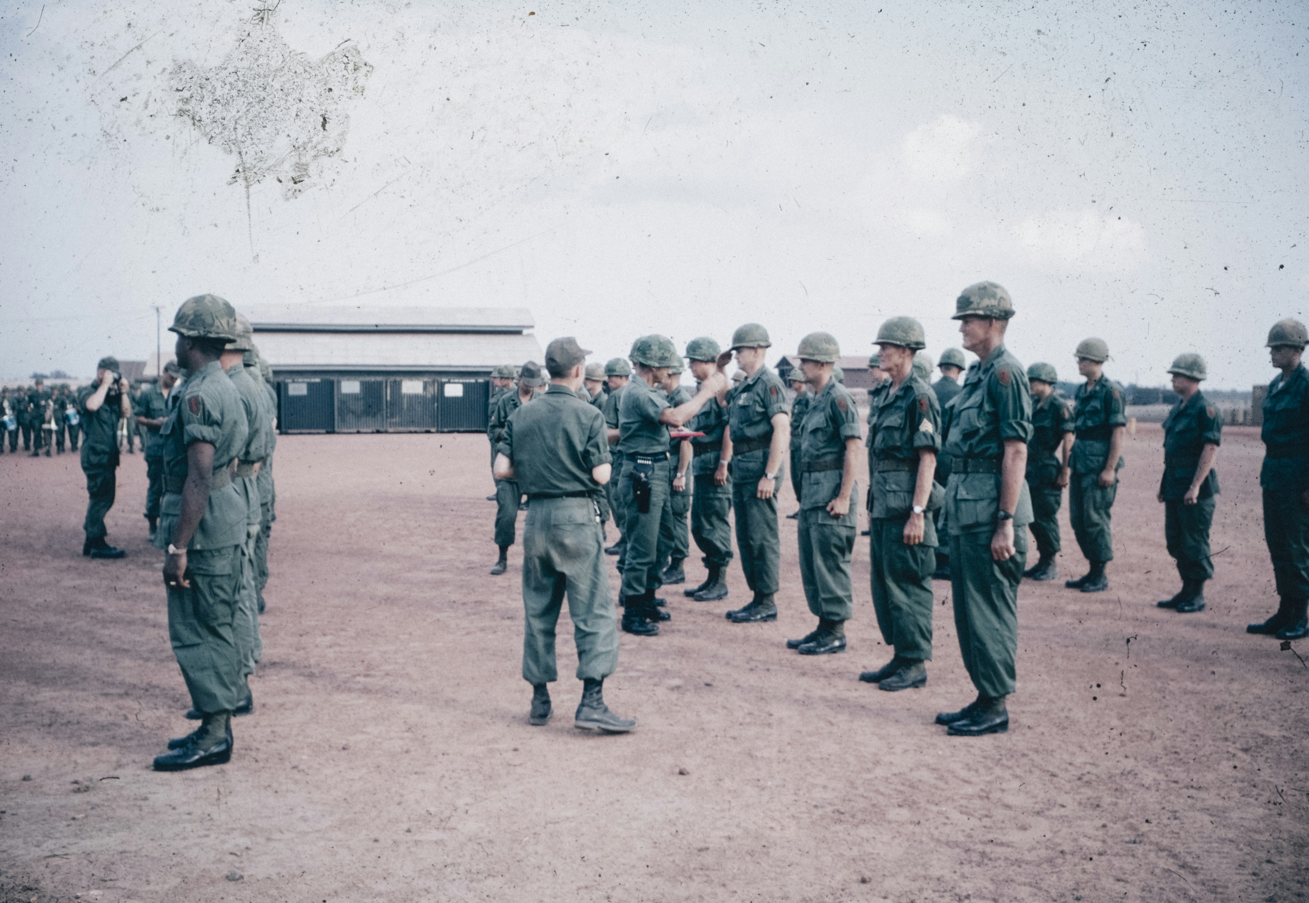 Military personnel in green uniforms and helmets standing in formation on a dirt field with buildings in the background.