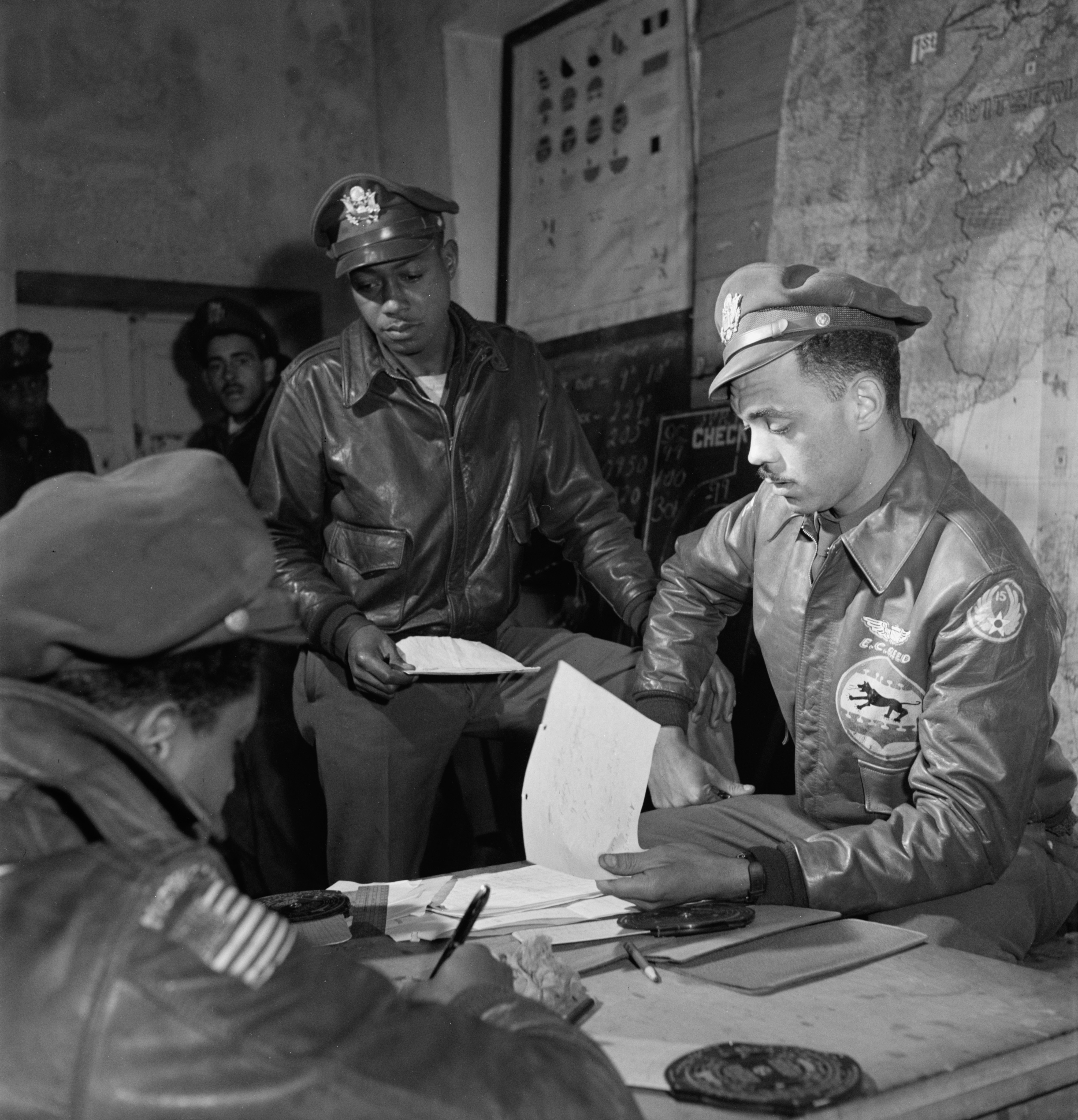 Three African American military officers in leather jackets are gathered around a table with papers, in a room with maps and charts on the walls.