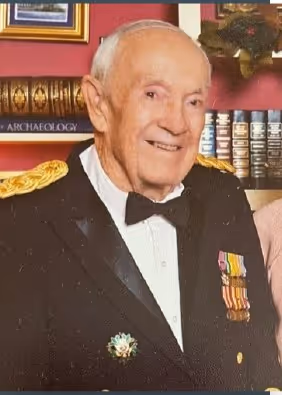 Elderly man wearing a formal black jacket with military-style medals and gold epaulettes, smiling in a room with books and framed pictures.
