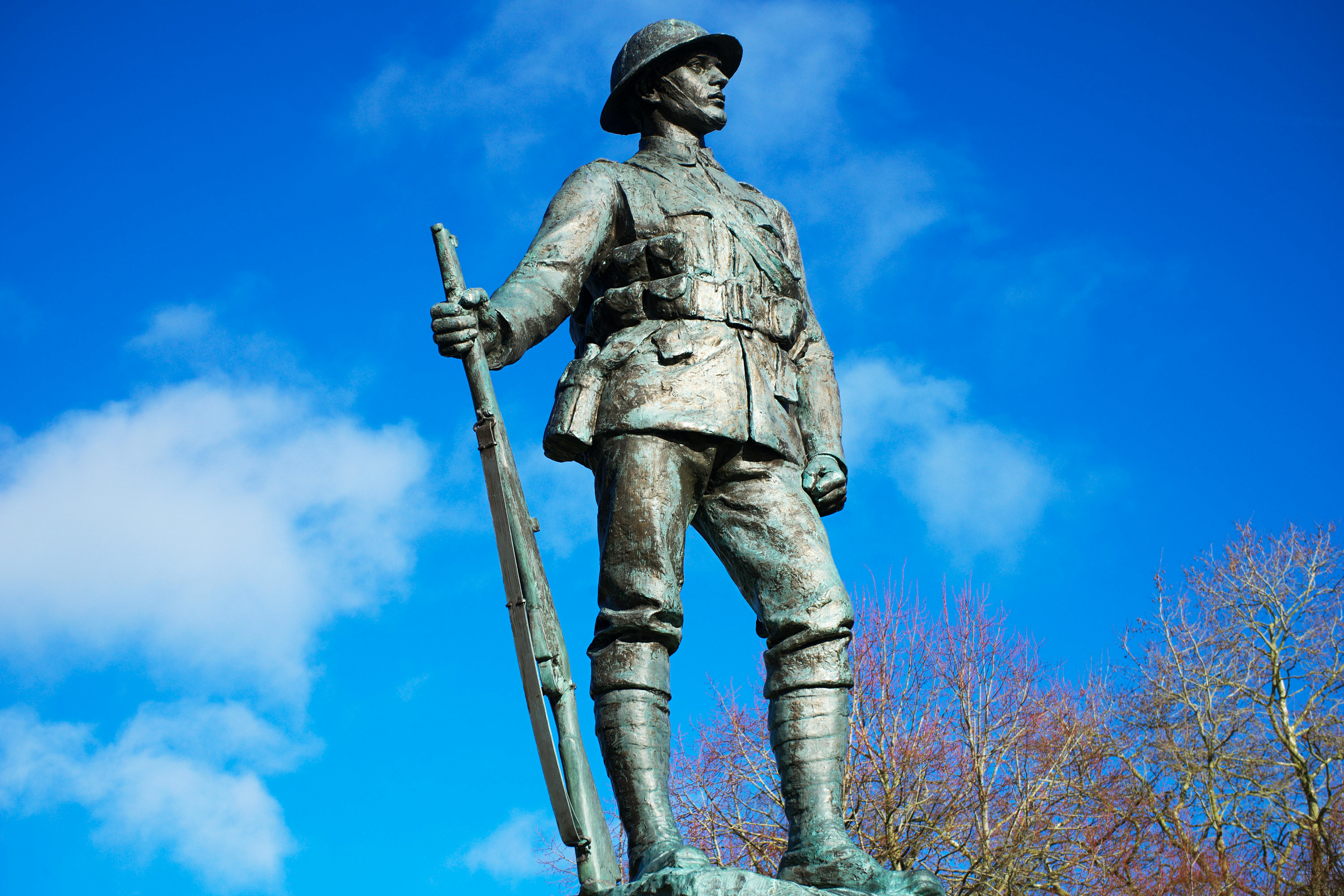 Bronze soldier statue in World War I military uniform holding a rifle against a blue sky with sparse clouds.