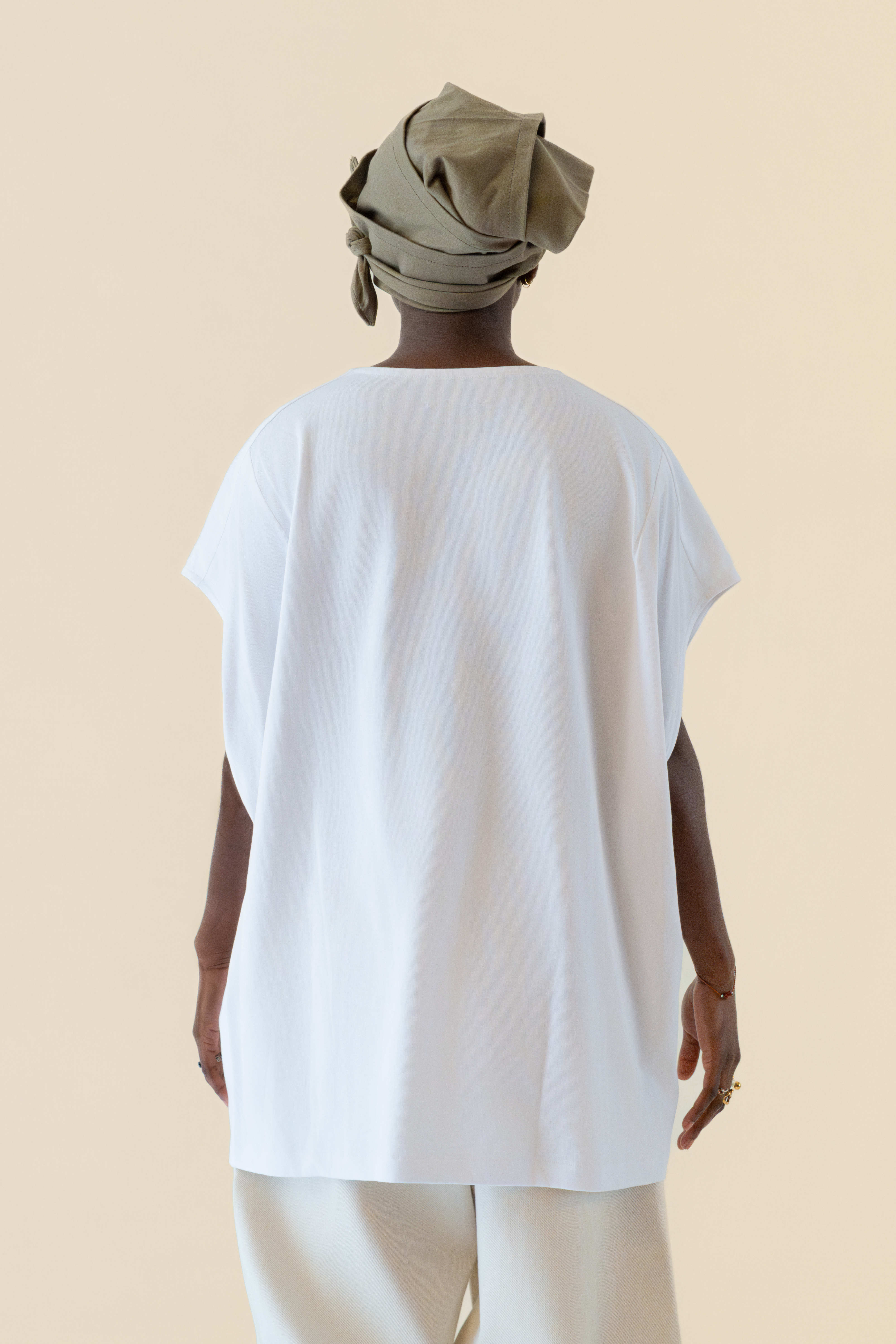 Person with dark skin wearing an olive green headwrap, white loose short-sleeve shirt, and light pants, seen from the back.