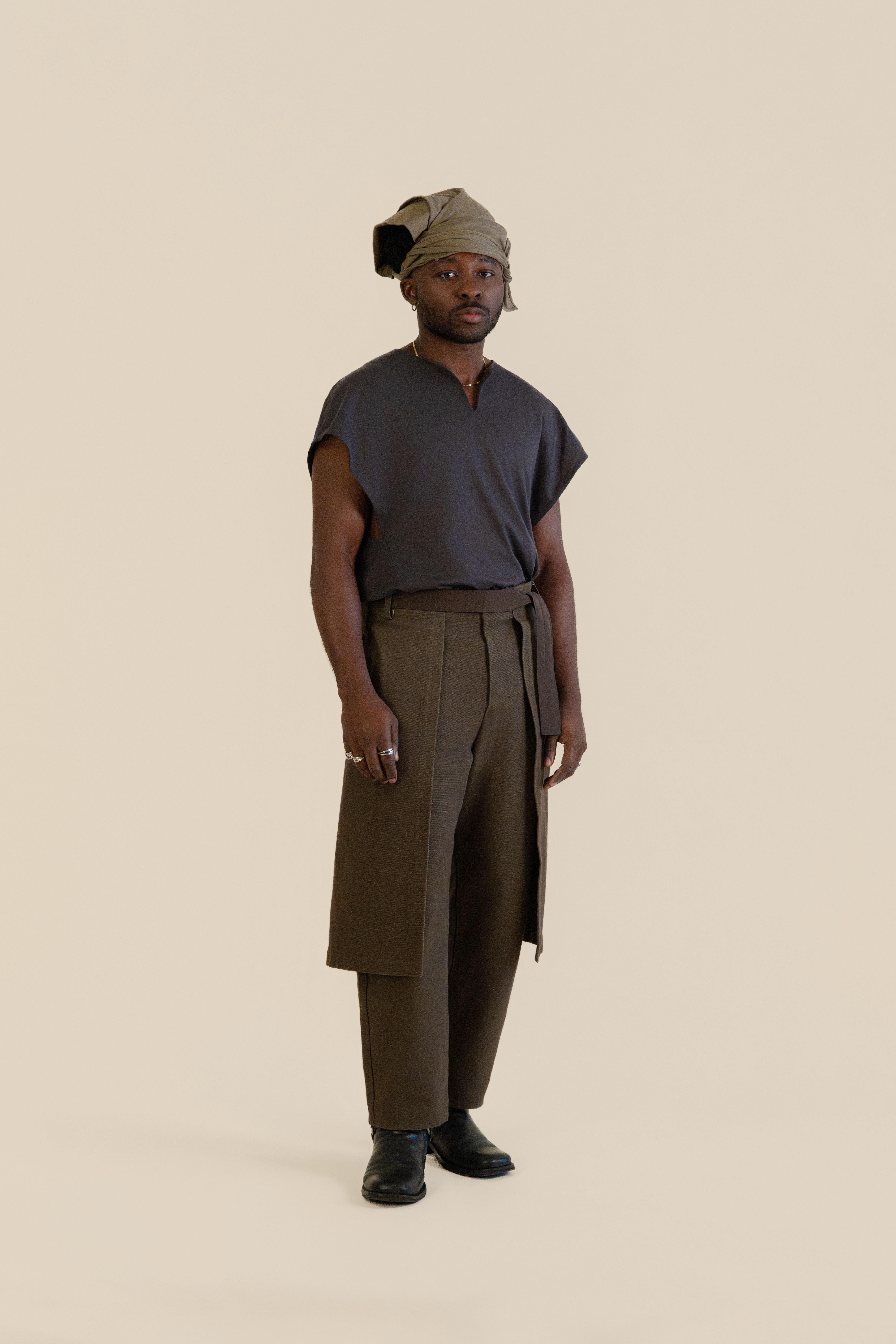 Full-body front view of male model wearing grey Light U-neck Tunic, taupe twisted bandana and brown trousers in soft warm light, Oniri editorial aesthetic