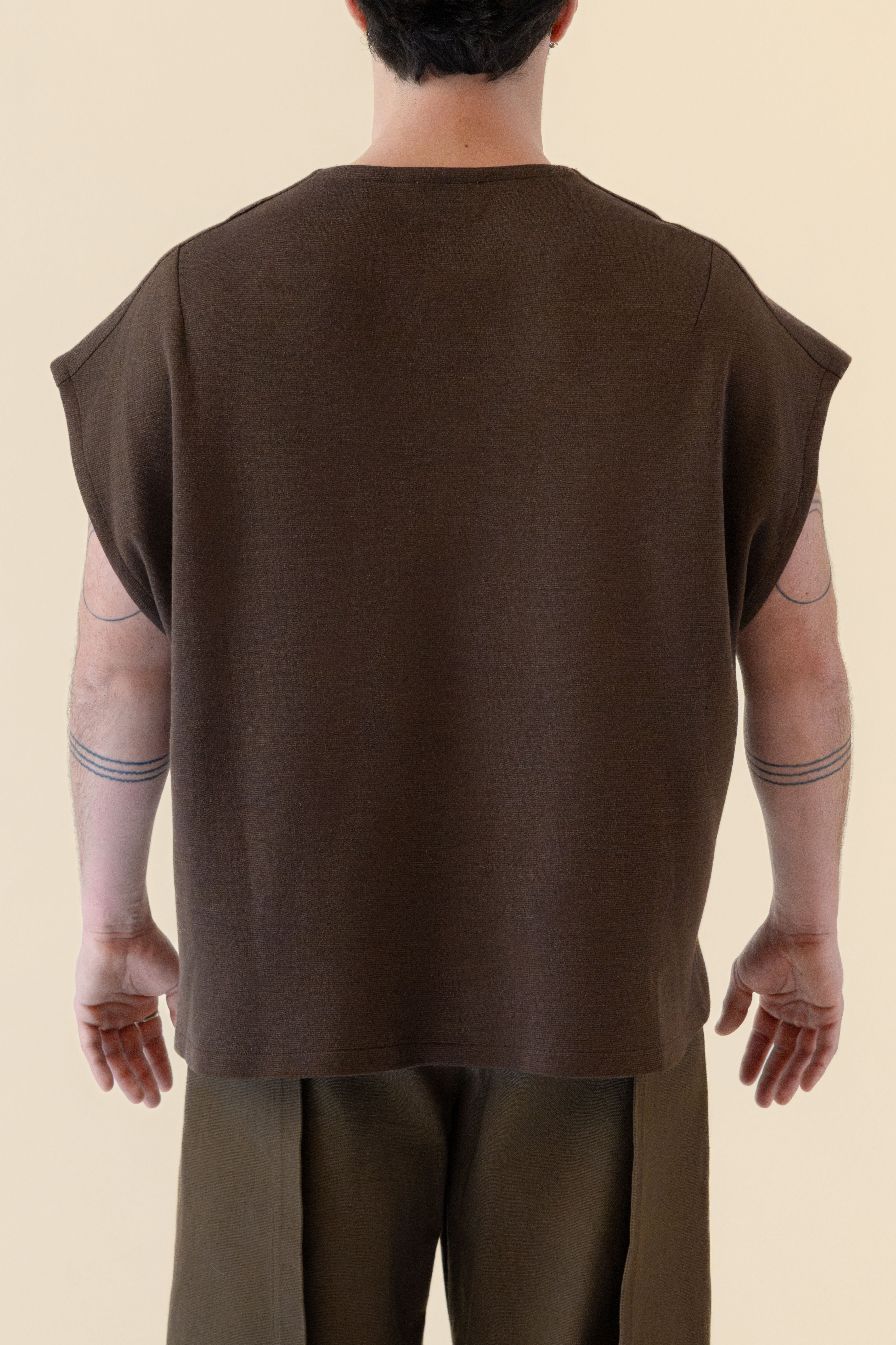 Back view of a person wearing a loose brown sleeveless top and matching pants, with tattoos visible on both arms.
