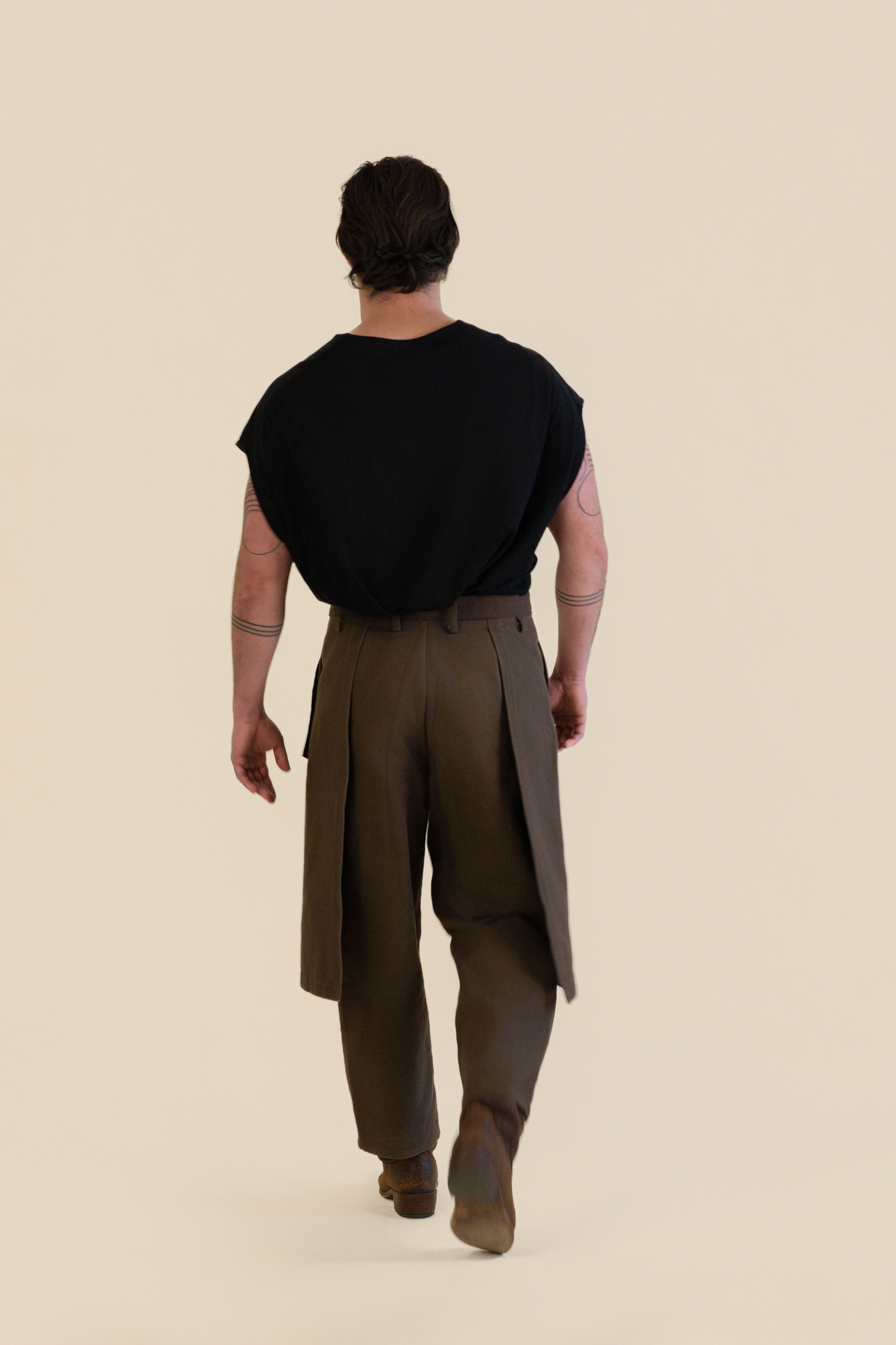 Man with tattoos on arms wearing a black sleeveless top and brown pants walking away against a beige background.