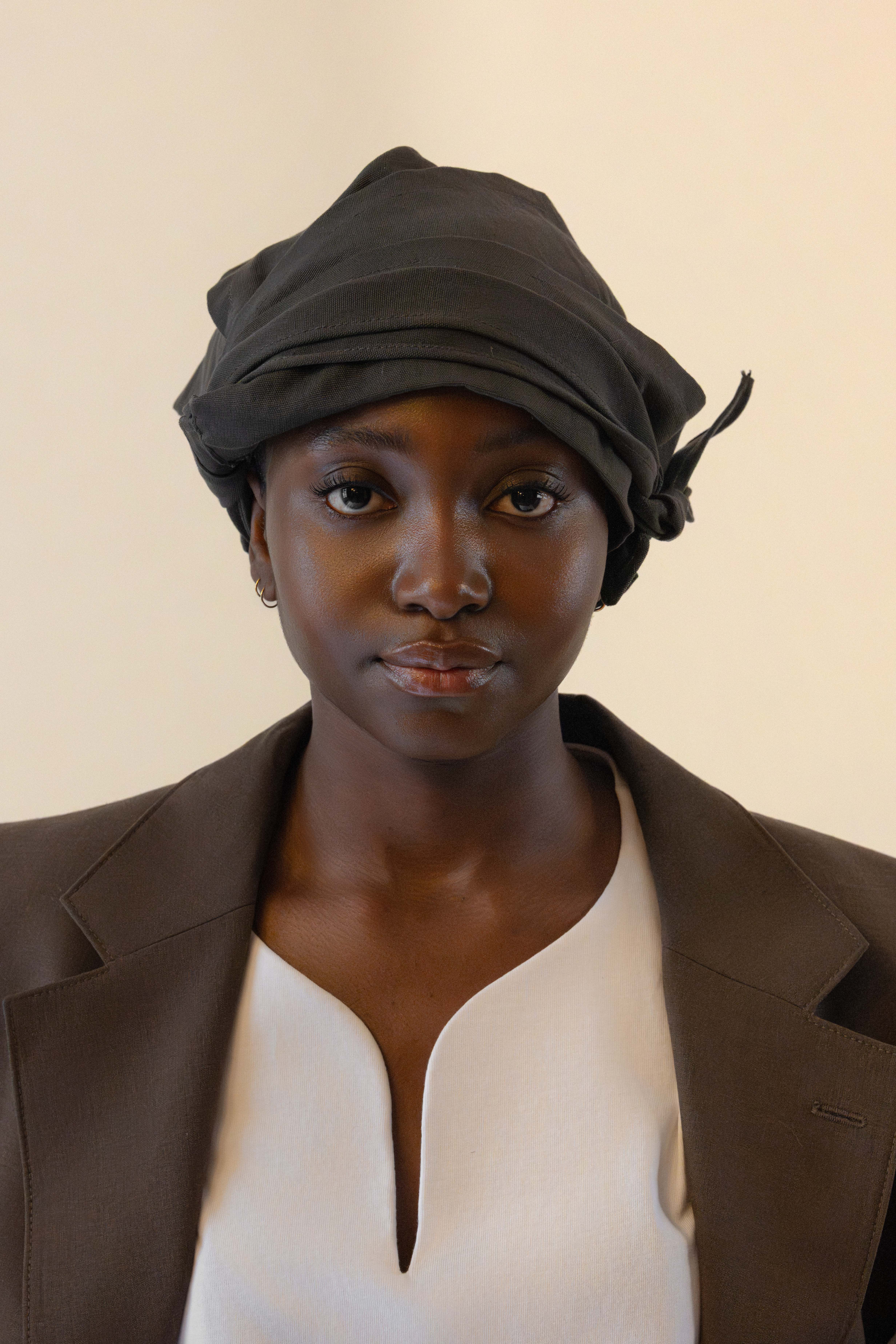 Portrait of a woman wearing a black head wrap, brown blazer, and white top against a beige background.