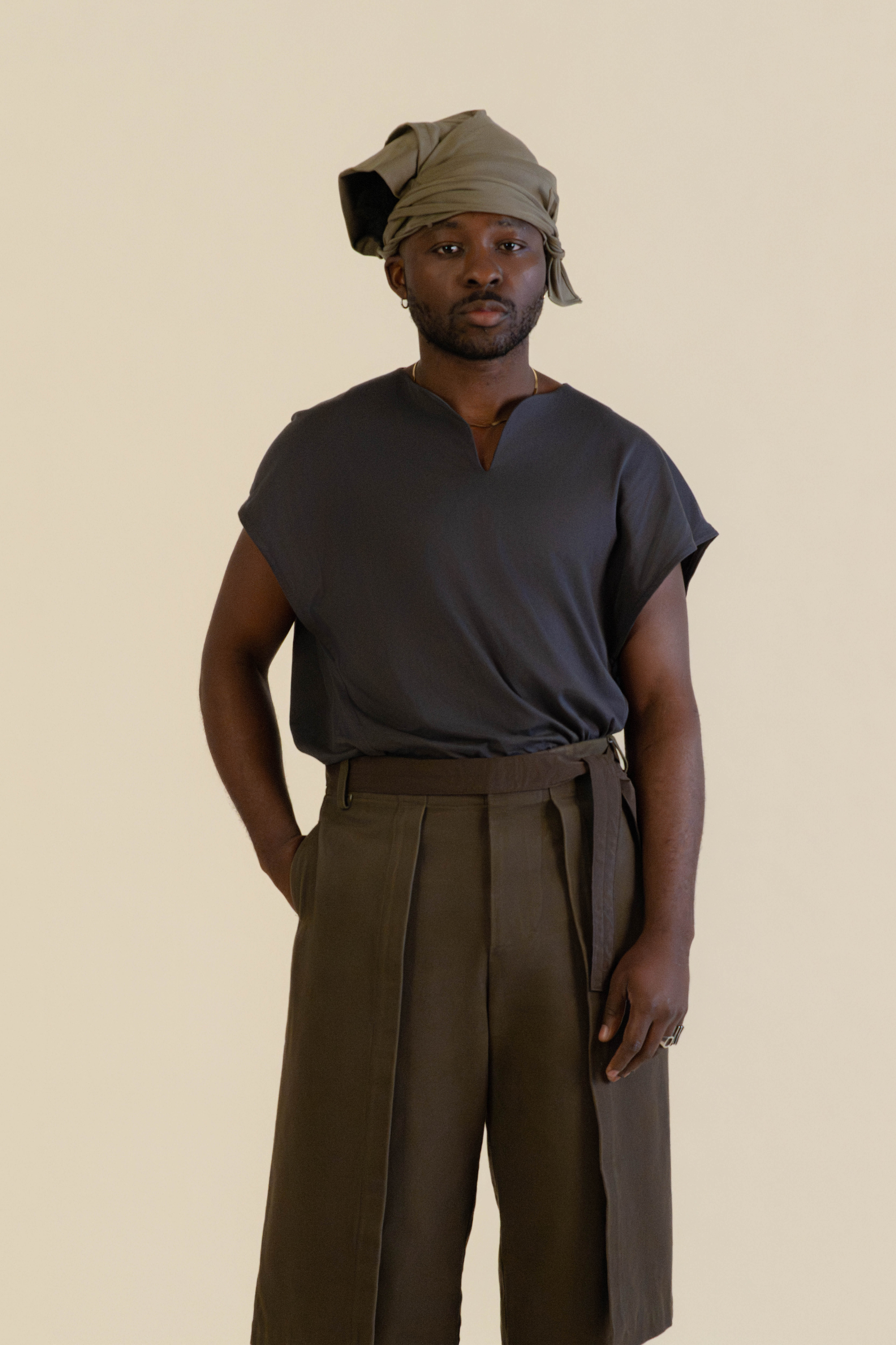 Front half-body view of male model wearing grey Light U-neck Tunic, taupe twisted bandana and brown trousers in soft warm light, Oniri editorial aesthetic