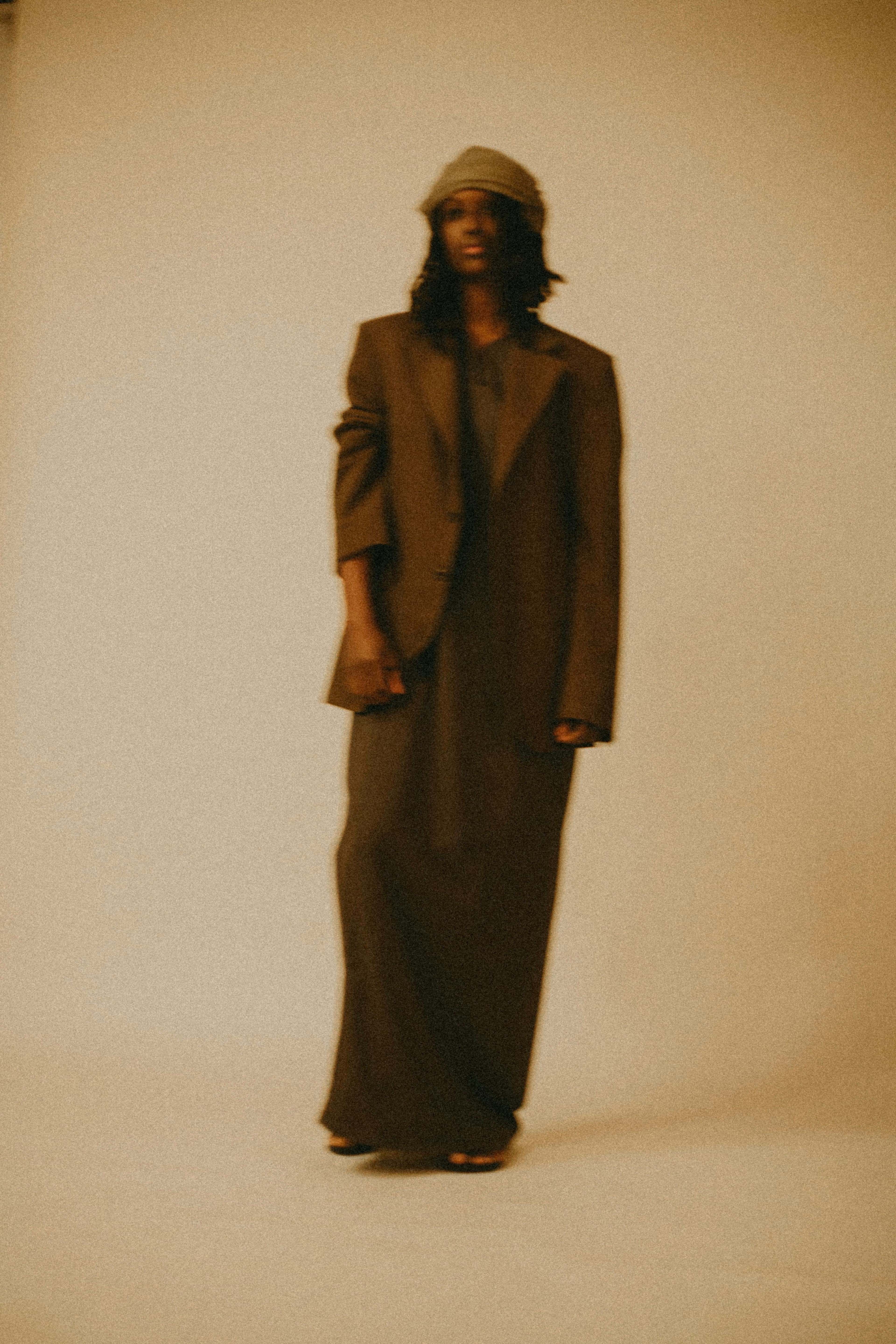 Model wearing an oversized dark brown blazer and matching maxi skirt, styled with a grey cap, standing against a neutral beige background.