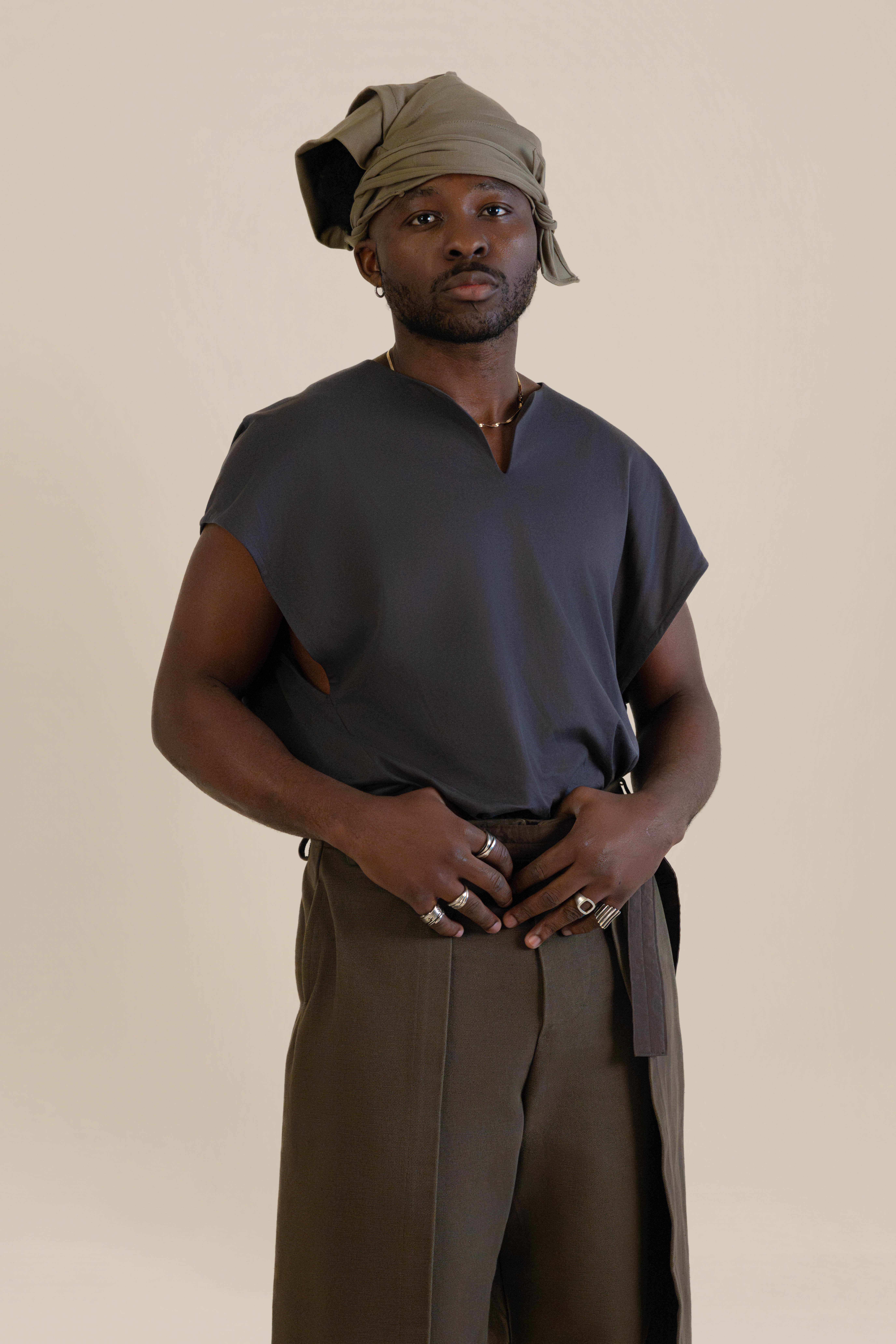 Front view of male model wearing grey Light U-neck Tunic, taupe twisted bandana and brown trousers in soft warm light, Oniri editorial aesthetic