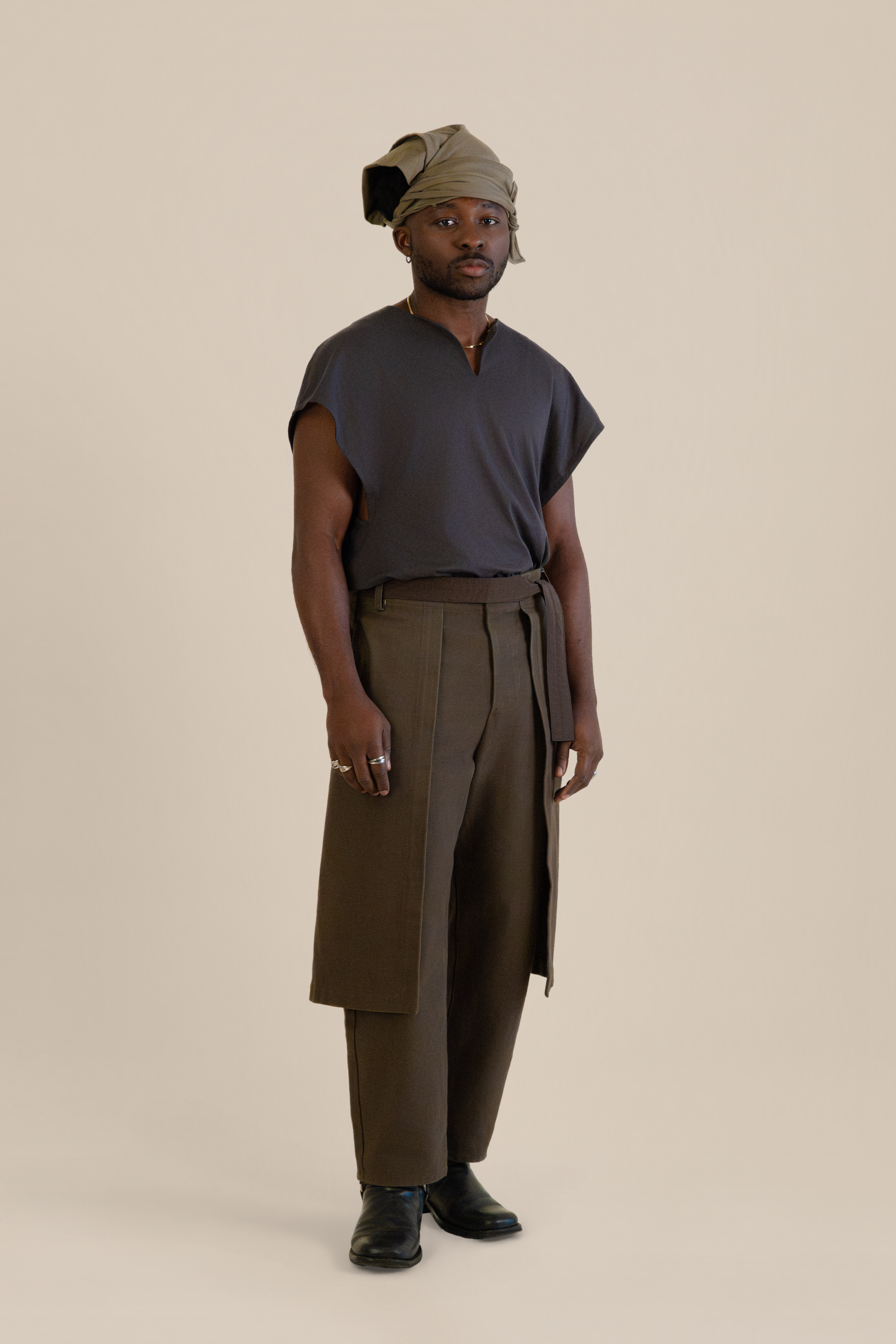 Full-body front view of male model wearing grey Light U-neck Tunic, taupe twisted bandana and brown trousers in soft warm light, Oniri editorial aesthetic