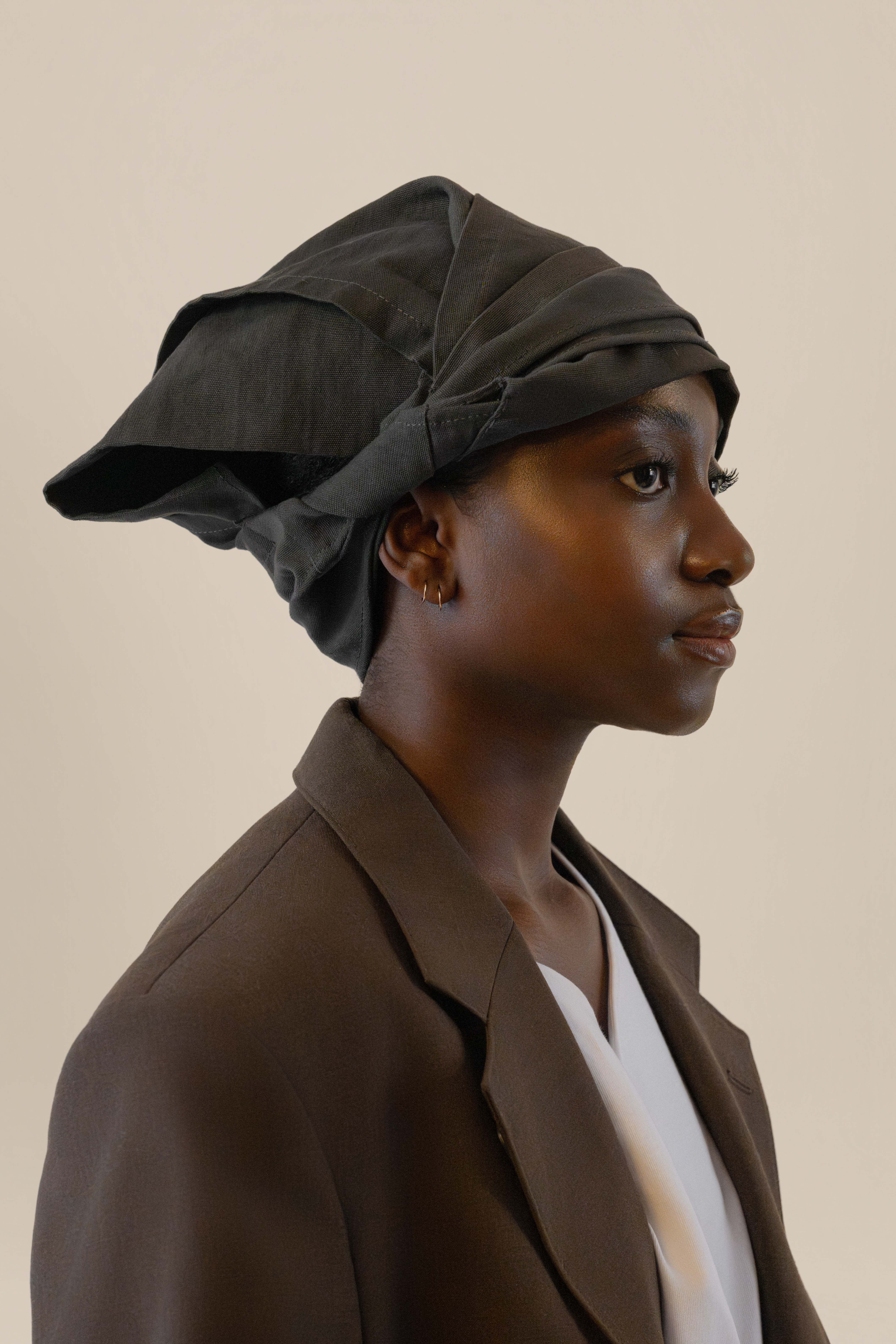 Side profile of female model wearing grey twisted bandana and brown blazer in soft warm light, Oniri editorial aesthetic