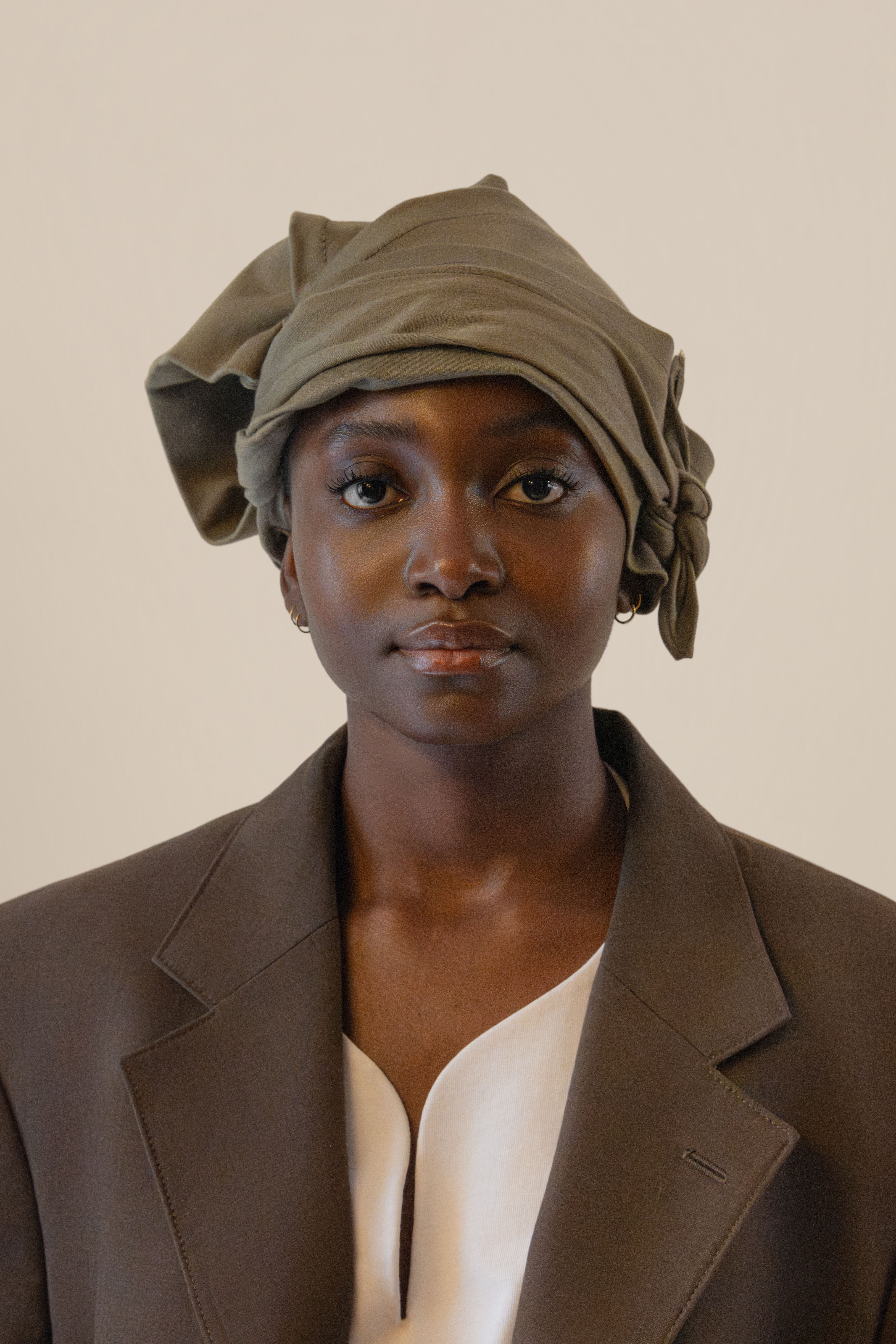 Frontal portrait of female model wearing grey twisted bandana, brown blazer and white tunic in soft warm light, Oniri editorial aesthetic
