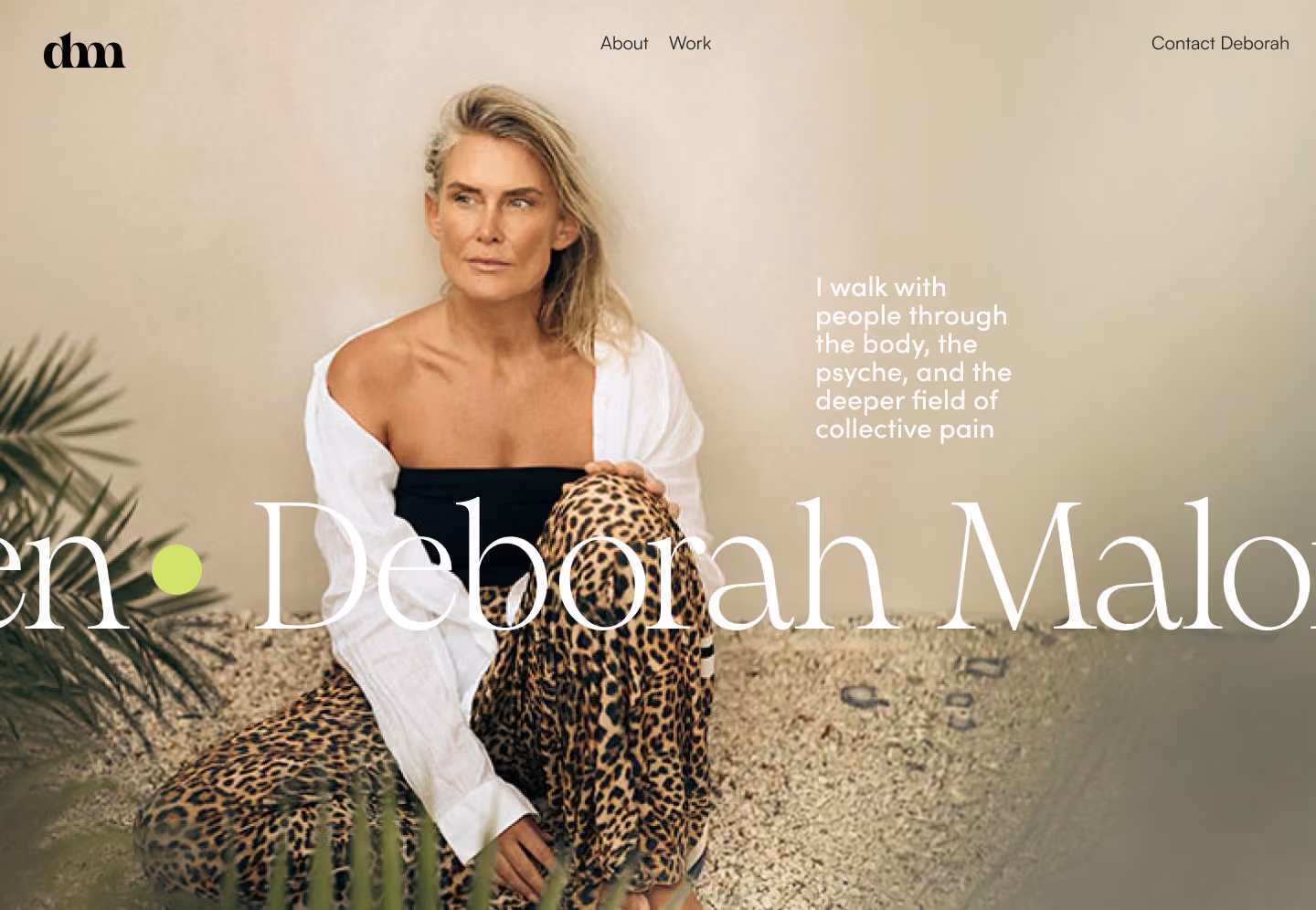 Woman with blonde hair in leopard pants and a white shirt sitting on gravel floor near plants.