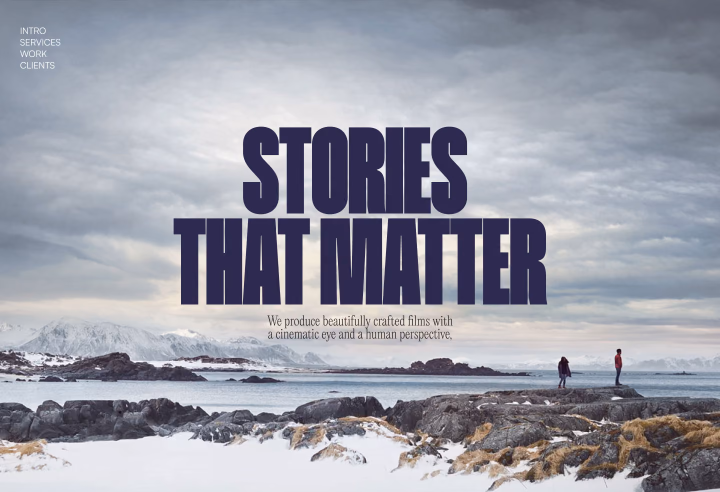 Two people on rocks by snowy shore with mountains and cloudy sky, text reads Stories That Matter.