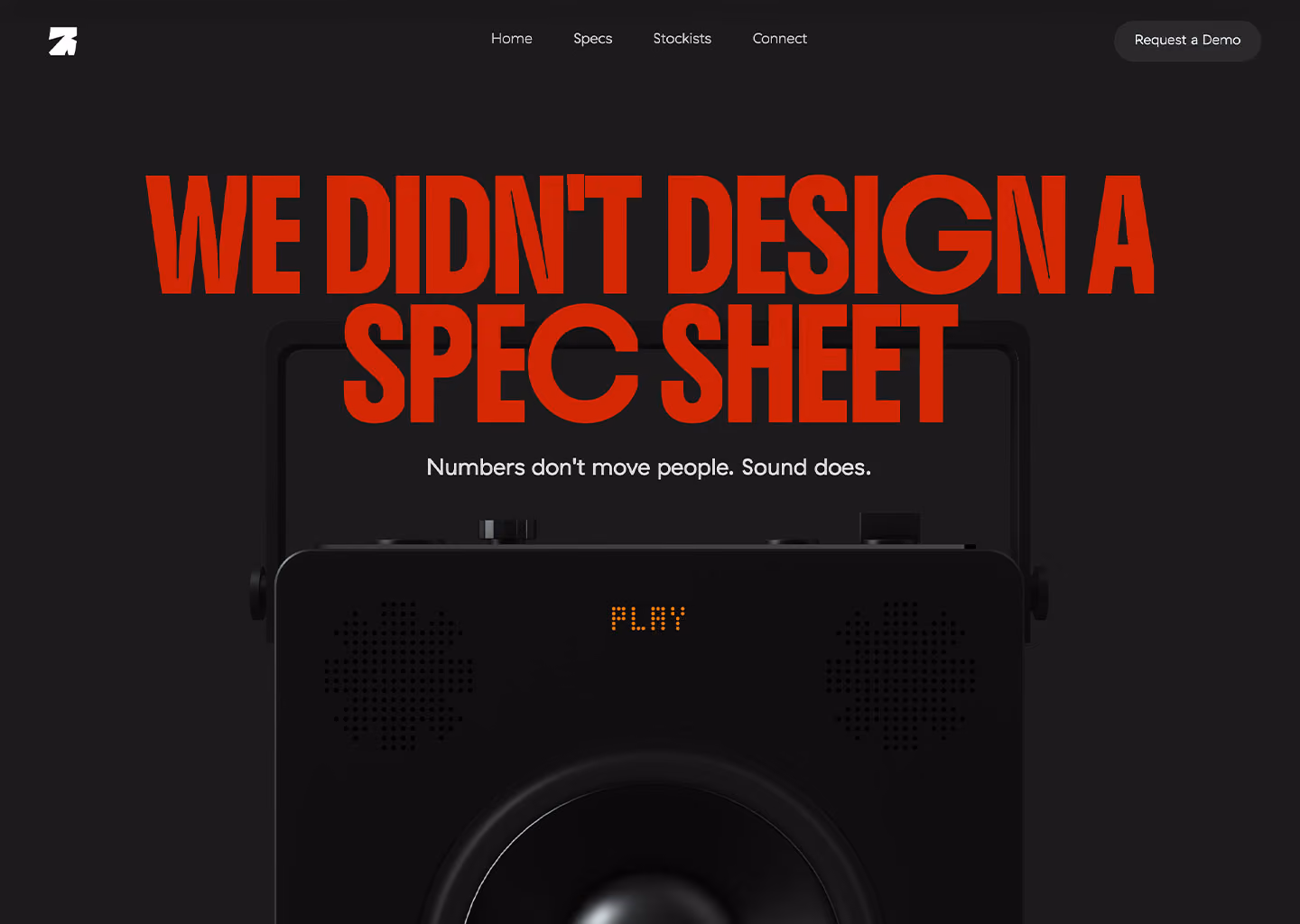 Black speaker with digital display reading 'PLAY' under bold red text 'WE DIDN'T DESIGN A SPEC SHEET' on dark background.