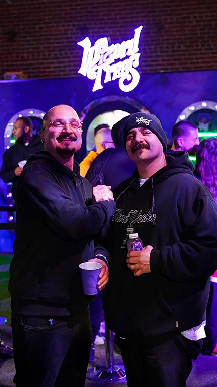 Two men in black hoodies smiling and shaking hands at a Wizard Trees event, with people and neon lights in the background.
