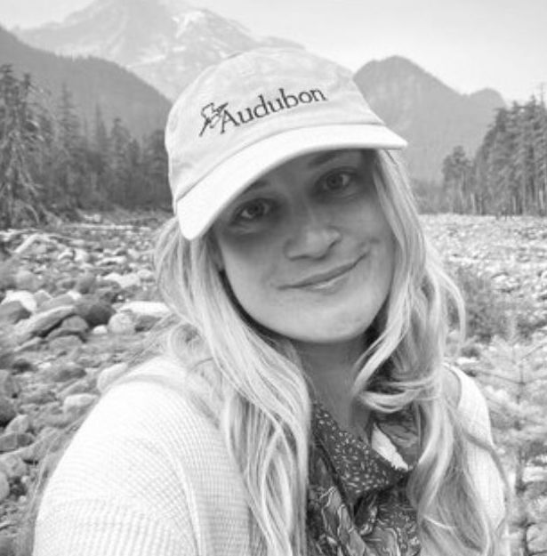 Smiling woman wearing an Audubon cap outdoors with mountains, rocks, and trees in the background.