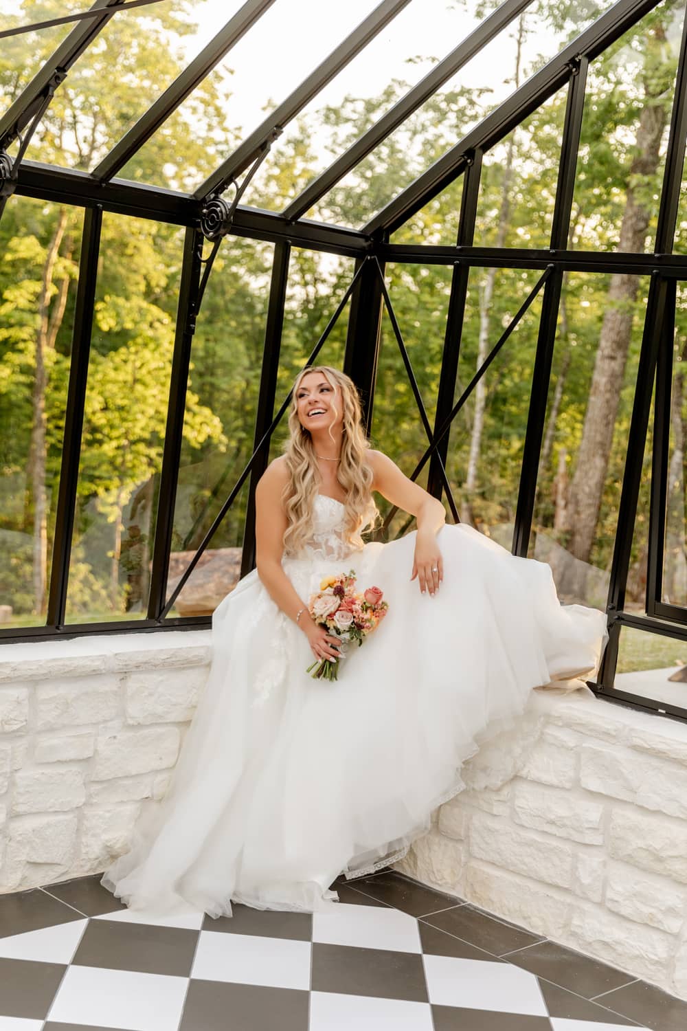 Bride in a flowing white gown sits in a glass greenhouse, surrounded by greenery and a checkered floor.