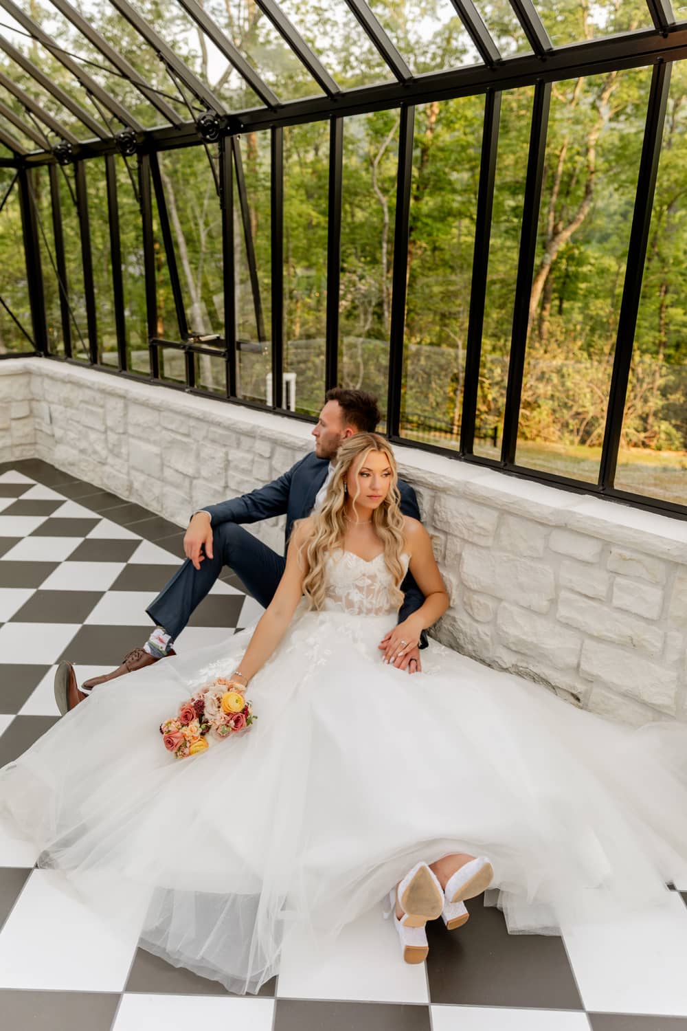 A couple sits on a checkered floor in a glass greenhouse, surrounded by greenery, celebrating their elopement.