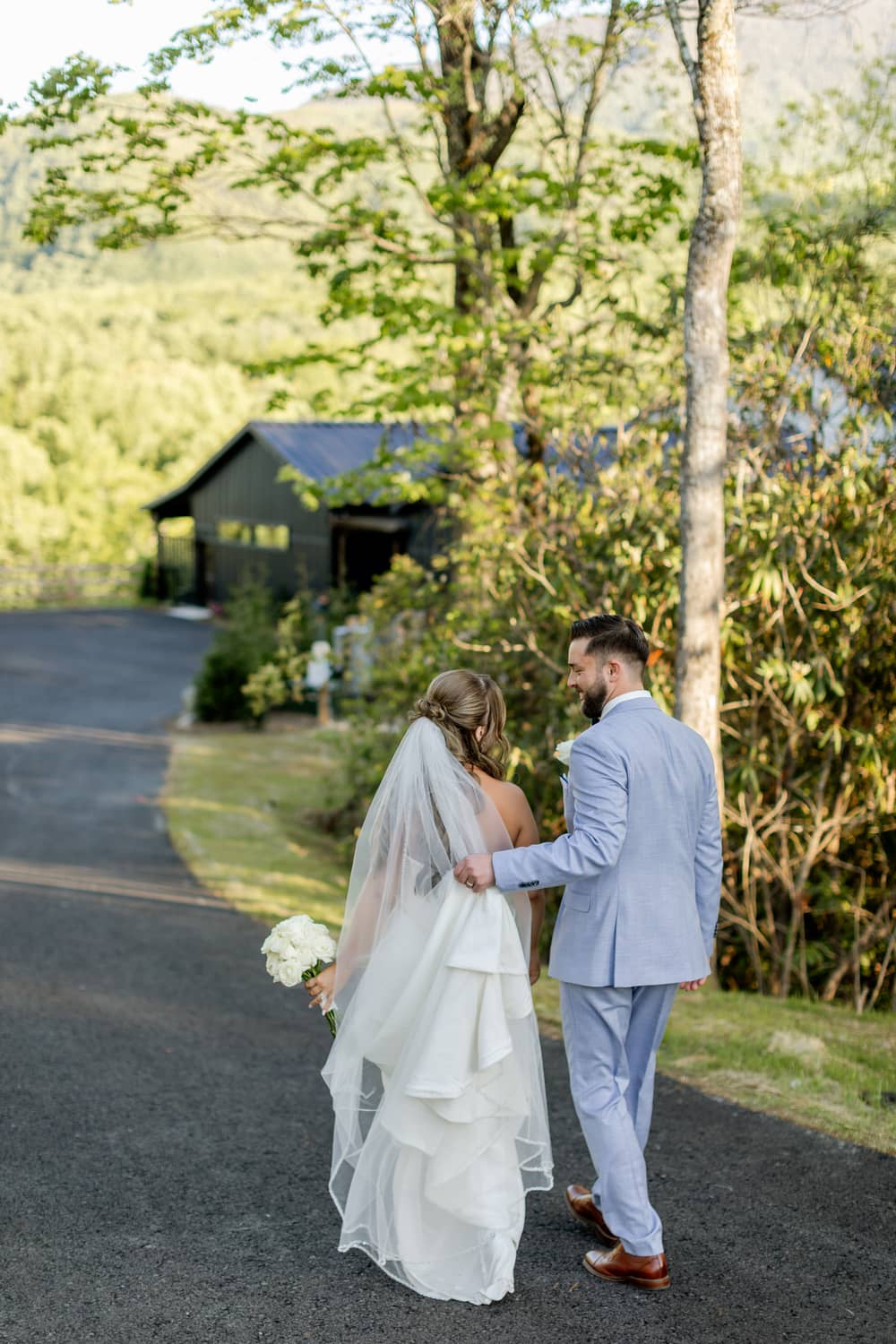 A couple in wedding attire stands on a scenic path, surrounded by trees, near a venue in the Smoky Mountains.