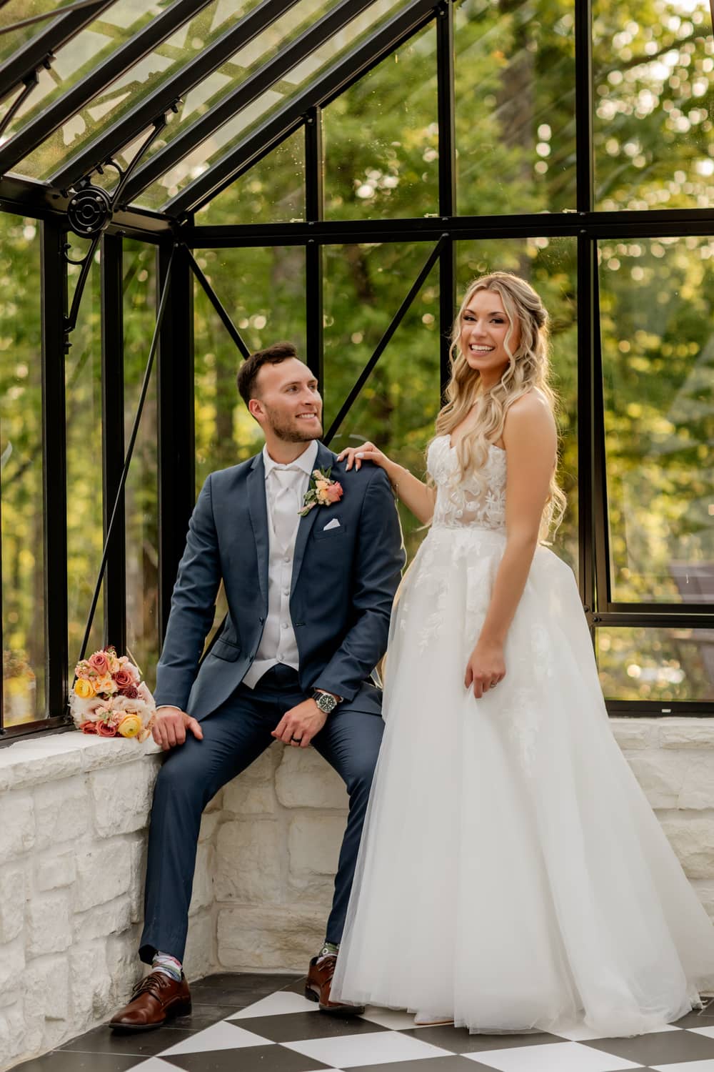 An elopement couple in wedding attire poses together in a bright, glass-enclosed greenhouse space surrounded by greenery.